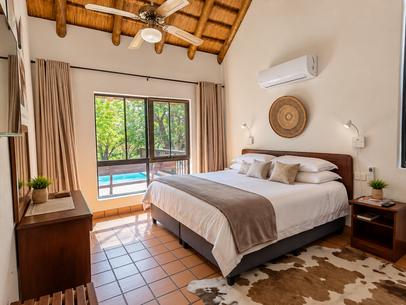 A well-maintained bedroom featuring a high thatched ceiling with exposed timber rafters, tiled flooring, and a split-system air conditioner, overlooking a pool area.