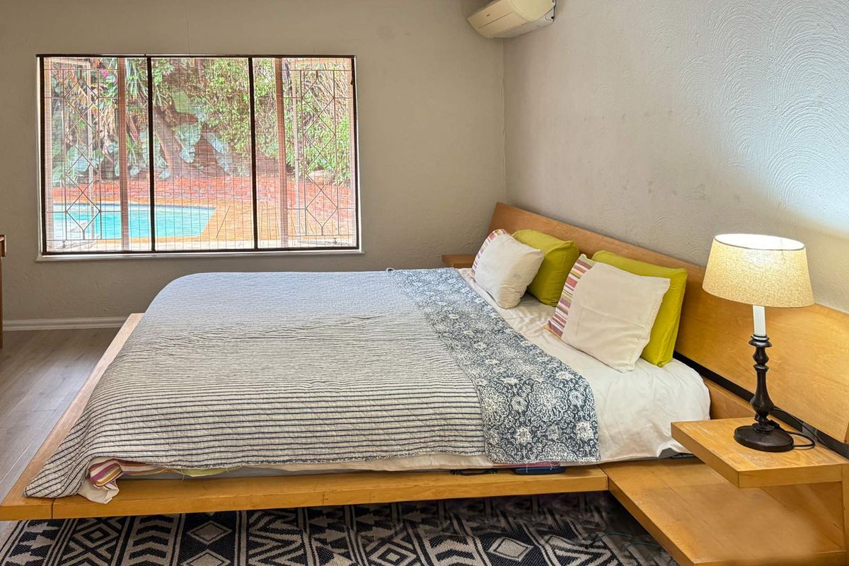 A bedroom featuring a large window with security bars, built-in headboard, and wall-mounted air conditioning unit, overlooking the outdoor pool area.