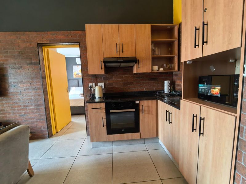 Compact kitchen featuring light wood cabinetry, granite countertops, built-in oven, extractor fan, and exposed brick accents, with direct access to the bedroom.