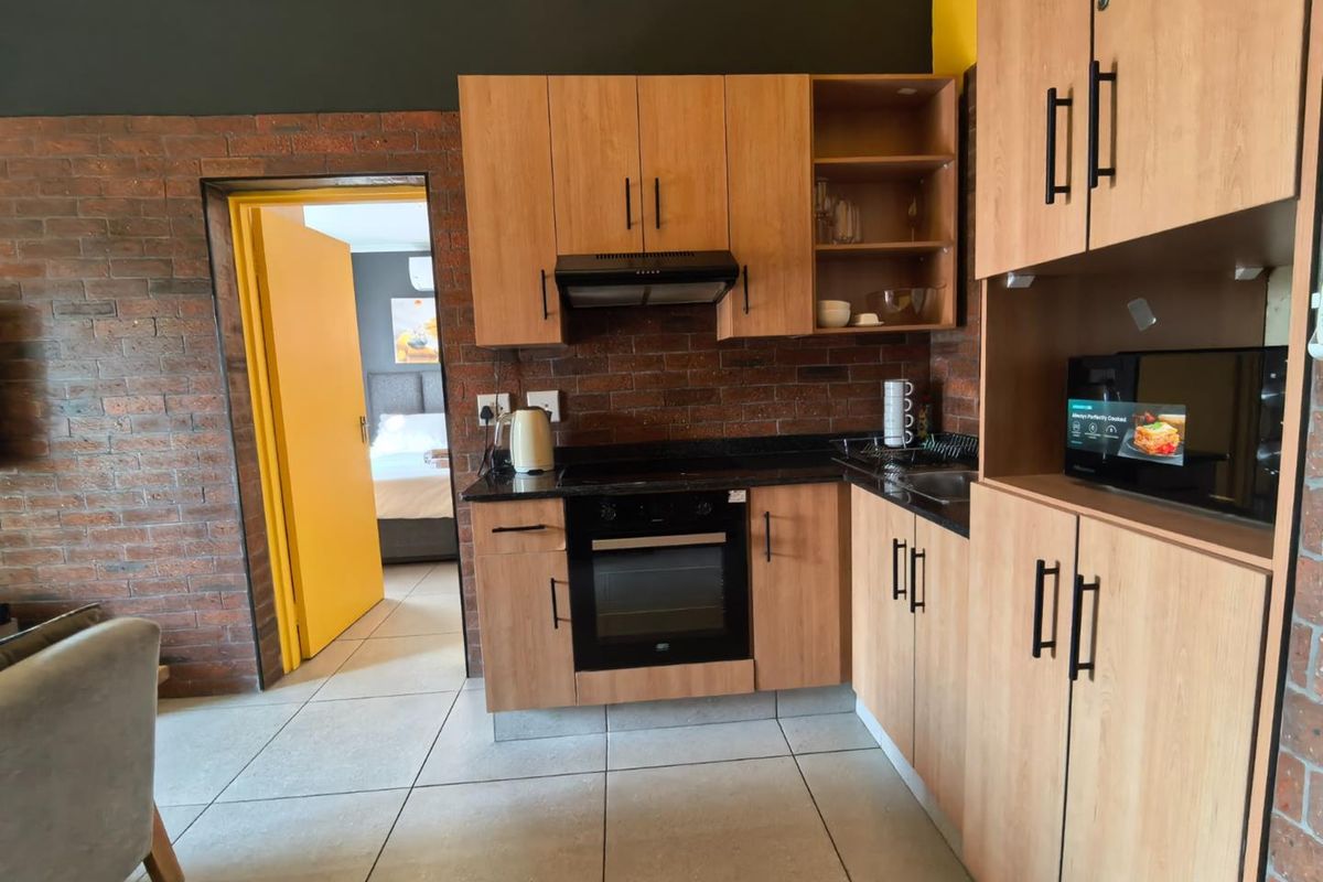 Compact kitchen featuring light wood cabinetry, granite countertops, built-in oven, extractor fan, and exposed brick accents, with direct access to the bedroom.