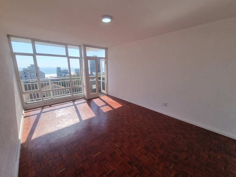 An empty living room featuring original parquet flooring and large glass doors providing natural light and access to a balcony with ocean views.