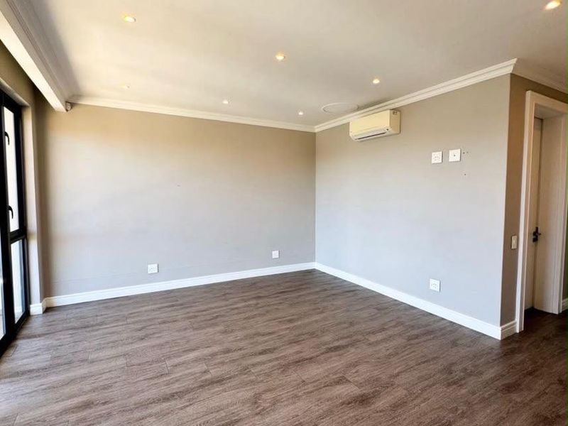 A bright, empty living room featuring modern laminate flooring, recessed downlighting, coving, and a wall-mounted split-system air conditioner.