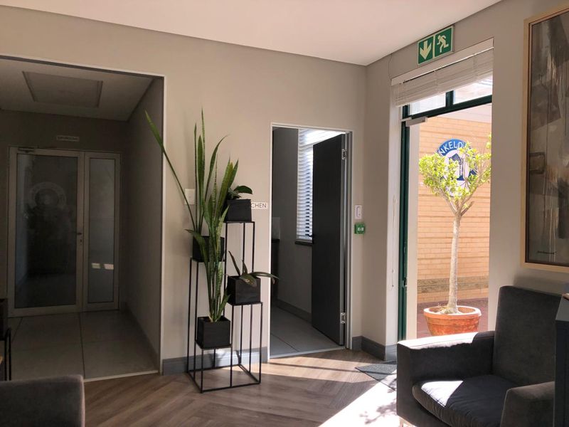 A bright office hallway featuring neutral walls, herringbone-style flooring, and access to adjacent rooms including a kitchen area.