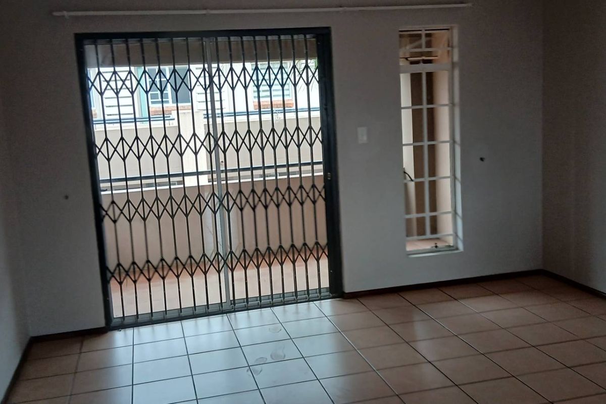 An empty living room featuring tiled flooring, a sliding glass door with a security gate, and a fixed window providing natural light.