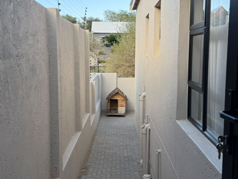 A narrow paved side passage featuring a boundary wall with electric fencing and an external air conditioning unit.
