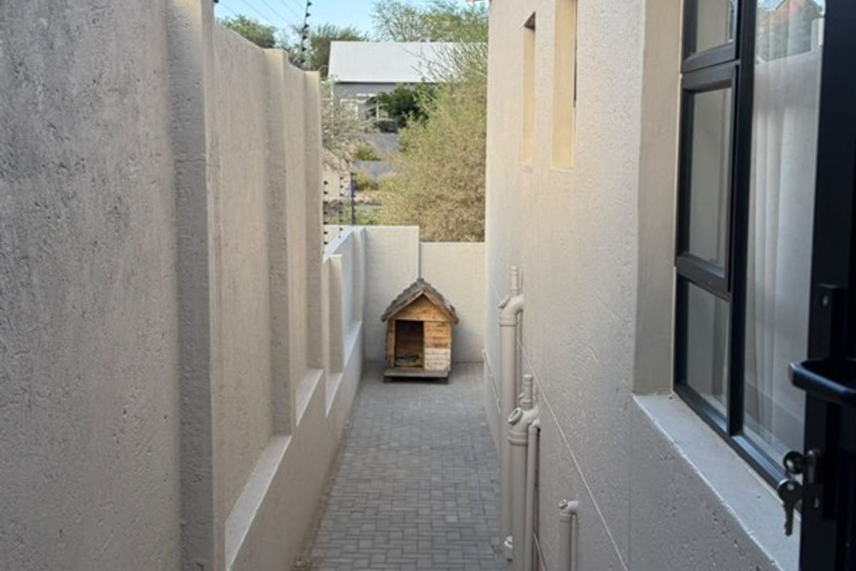 A narrow paved side passage featuring a boundary wall with electric fencing and an external air conditioning unit.