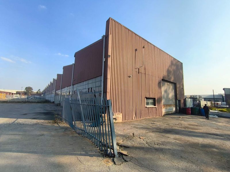 Industrial warehouse facility featuring corrugated metal cladding, a large roller shutter door, and an expansive asphalt yard area with perimeter security fencing.