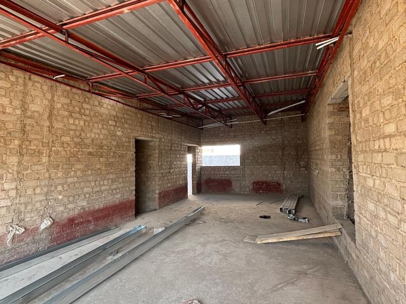 An unfinished interior space featuring exposed brick walls, a metal roof structure, and a concrete floor, currently in the construction phase.