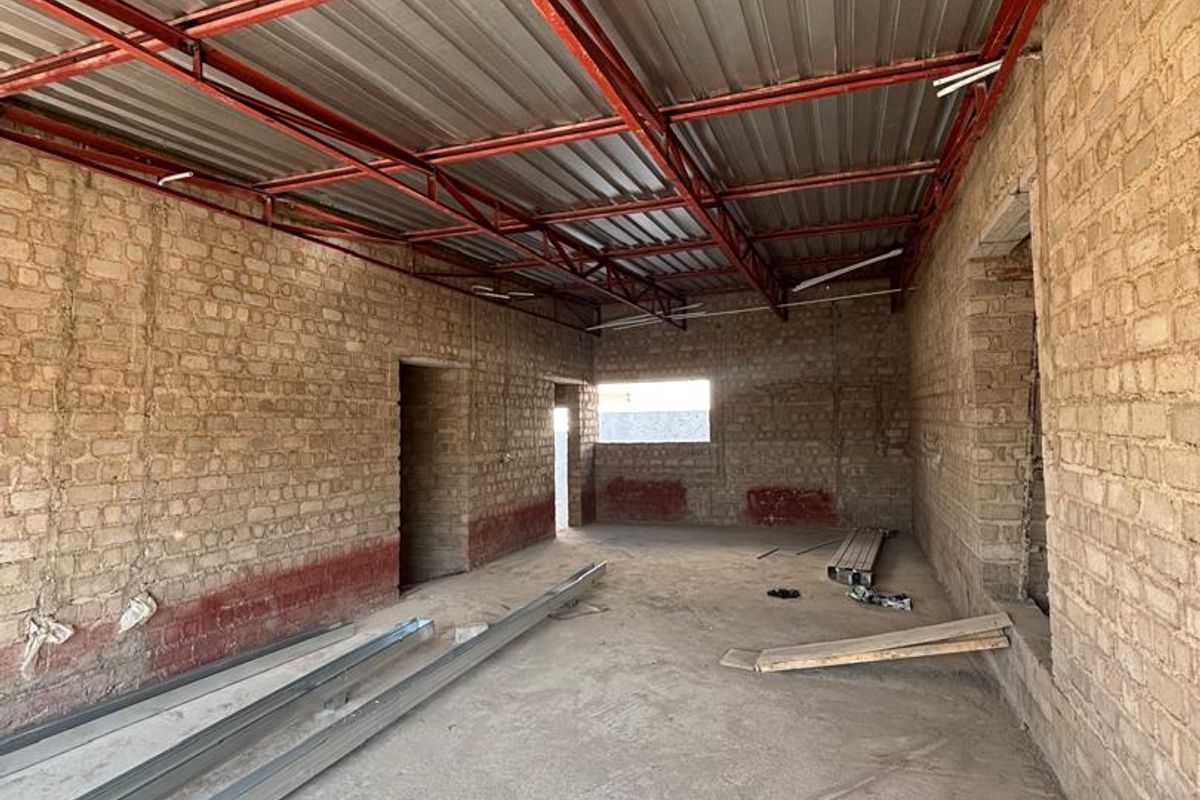 An unfinished interior space featuring exposed brick walls, a metal roof structure, and a concrete floor, currently in the construction phase.