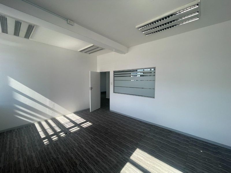 A bright, empty office space featuring modern carpet tiles, recessed fluorescent lighting, and a frosted glass partition window, offering a clean professional environment.