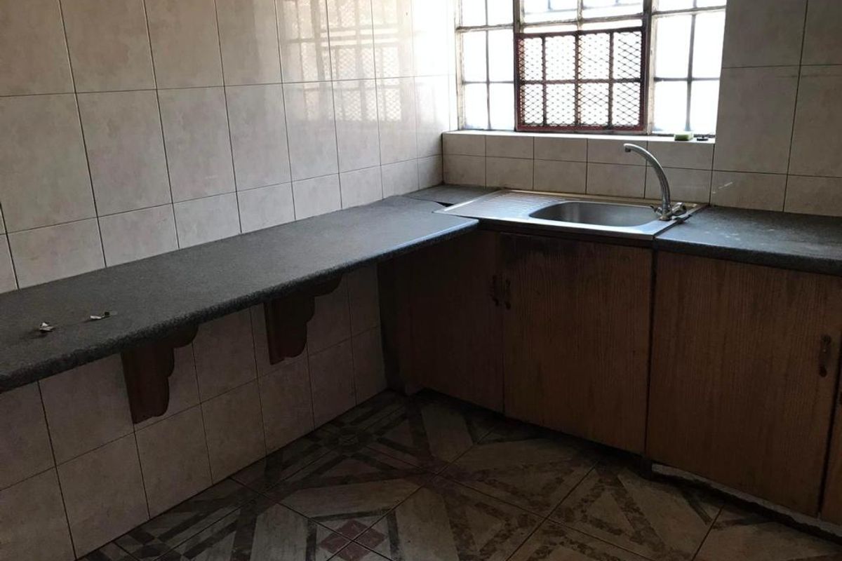 A dated kitchen area featuring a stainless steel sink, laminate countertops, tiled walls, and patterned floor tiles with a metal-framed window.