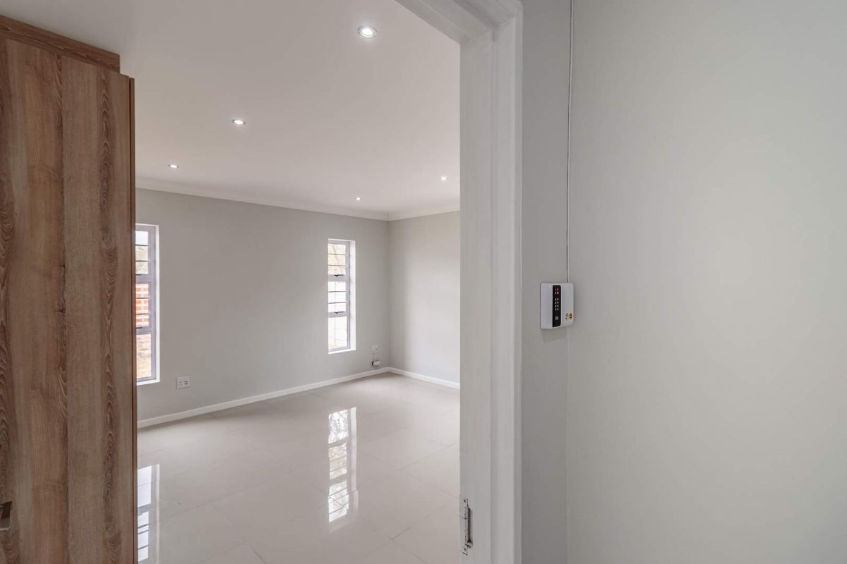 A bright, empty bedroom featuring neutral walls, polished tiled flooring, recessed downlighting, and an integrated alarm system keypad.