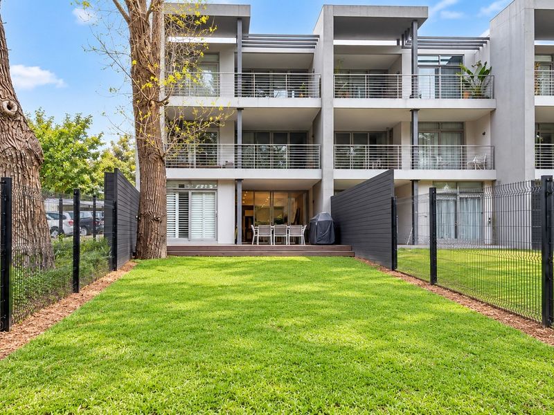 Rear view of a modern multi-story apartment complex featuring private ground-floor timber decking, a well-maintained lawn, and secure perimeter fencing.
