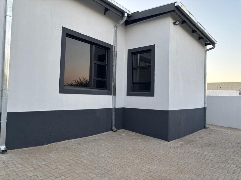 A well-maintained rear exterior view featuring modern white and dark grey painted walls, aluminium window frames, and a clean paved courtyard area.