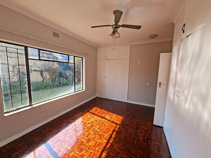 A bright, empty bedroom featuring classic parquet flooring, built-in storage cupboards, and a ceiling fan, with large windows overlooking the garden.
