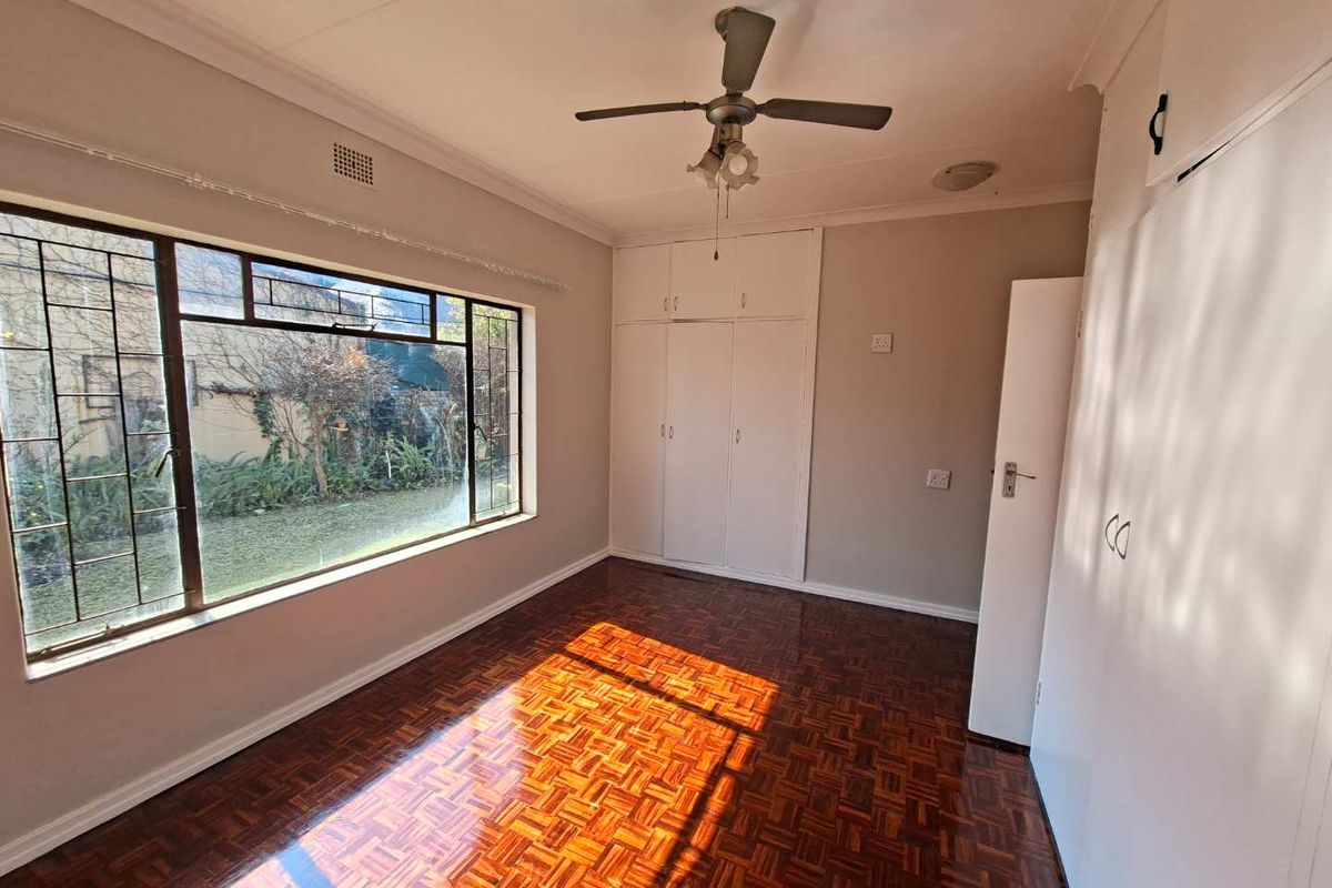 A bright, empty bedroom featuring classic parquet flooring, built-in storage cupboards, and a ceiling fan, with large windows overlooking the garden.