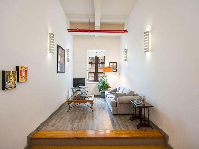 A narrow living room featuring high ceilings with exposed red piping, wall-mounted lighting, and wood-look laminate flooring with a timber step transition.