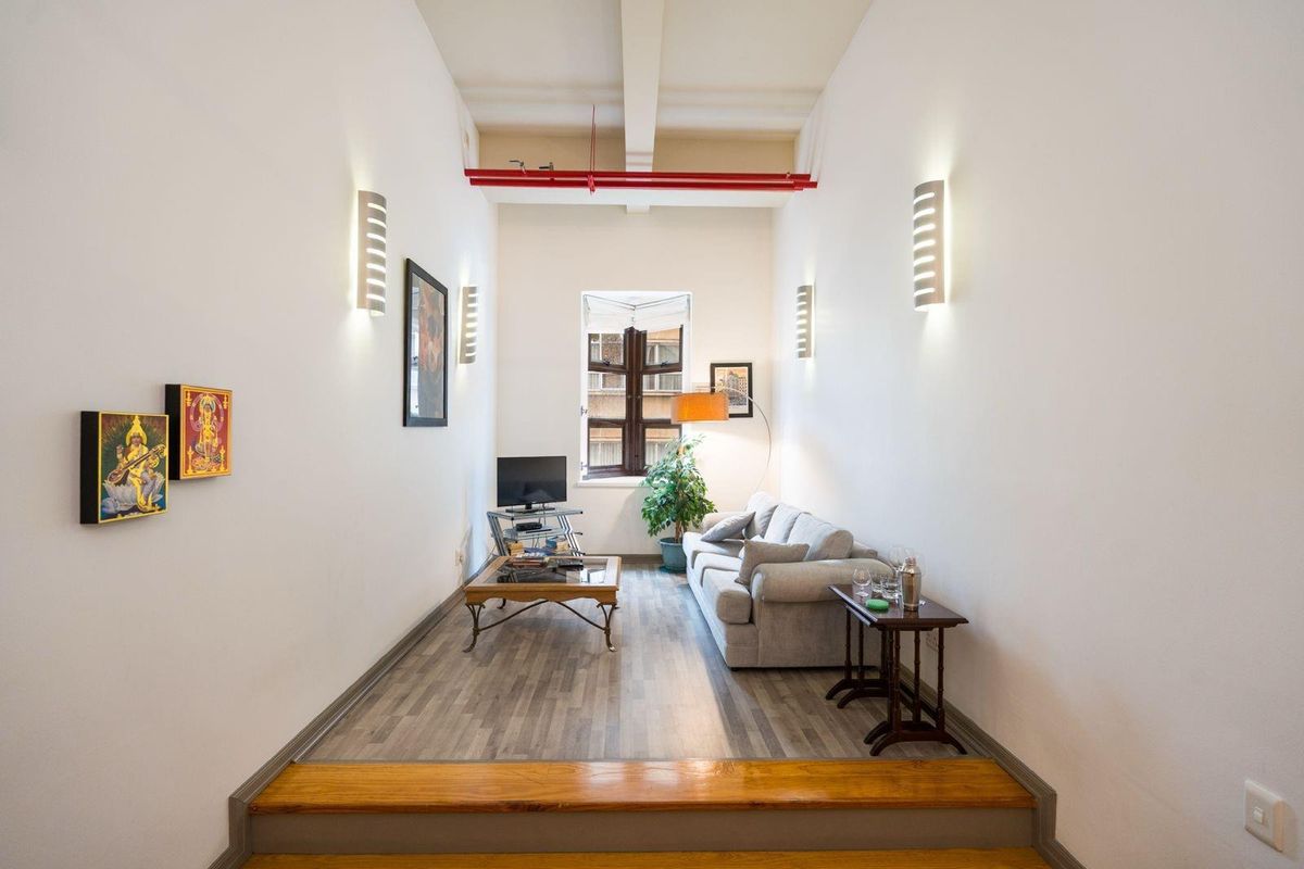 A narrow living room featuring high ceilings with exposed red piping, wall-mounted lighting, and wood-look laminate flooring with a timber step transition.