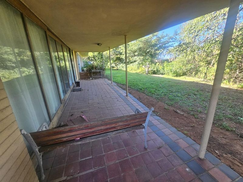 A covered outdoor patio area featuring paved flooring and large sliding glass doors, overlooking a spacious garden.