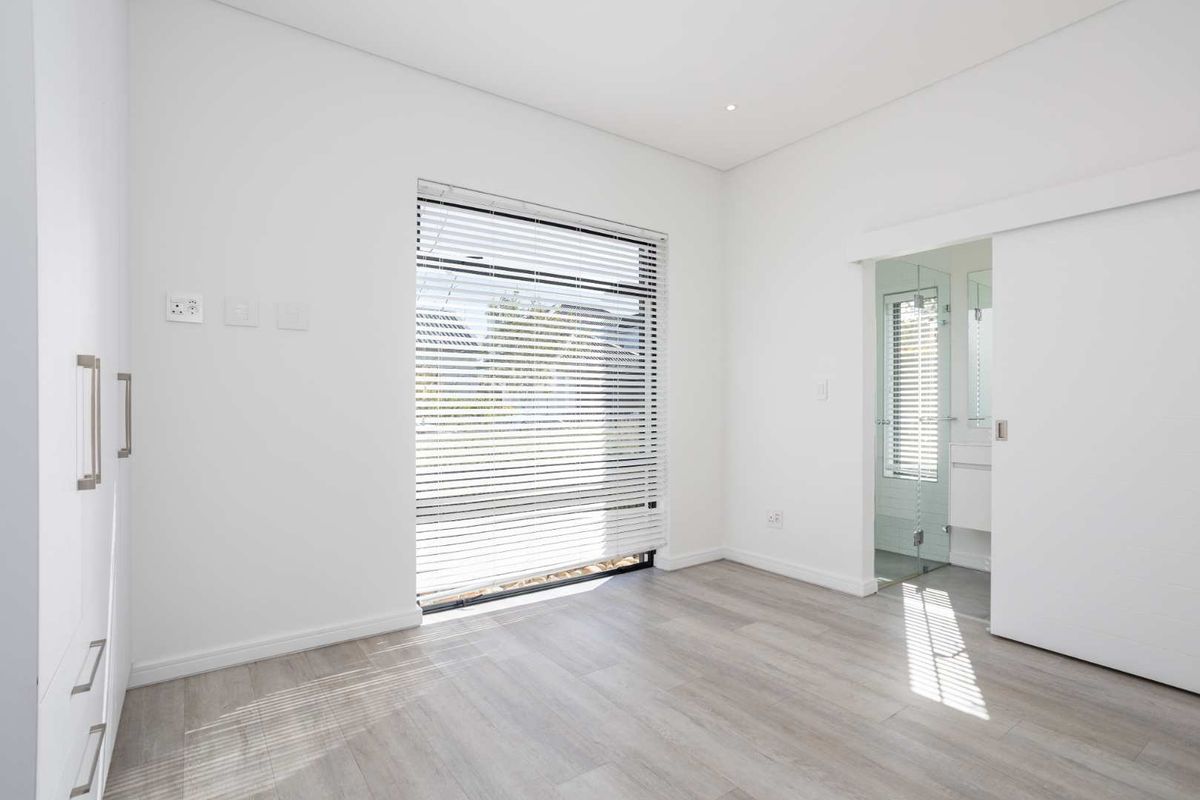 A bright, modern bedroom featuring neutral laminate flooring, built-in cupboards, and an en-suite bathroom accessed via a sliding door.