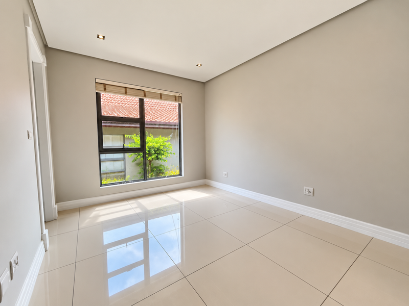 A bright, empty bedroom featuring neutral walls, polished tiled flooring, recessed downlights, and a large window with views of the exterior.