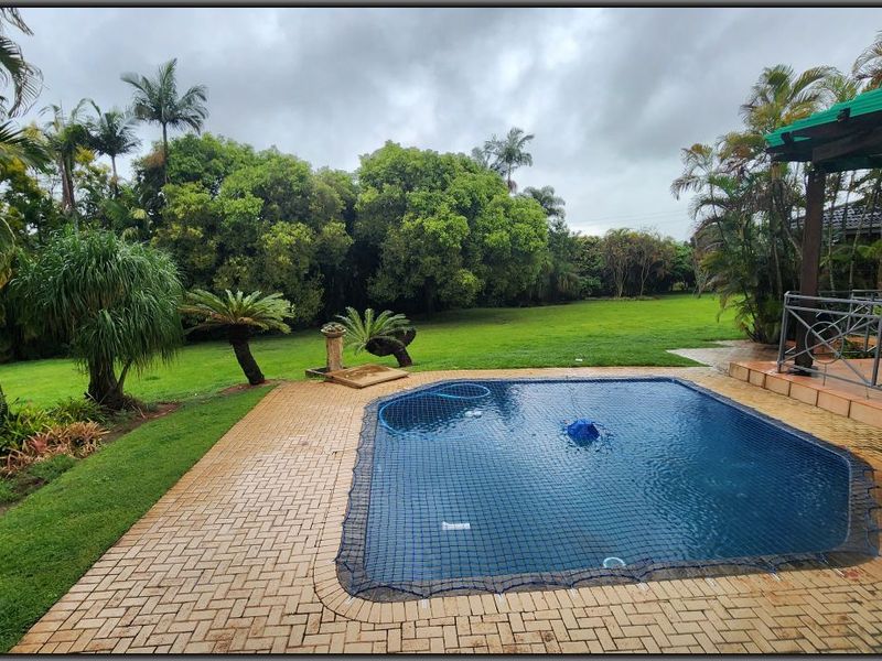 Outdoor pool area featuring a paved deck, safety net, and a covered patio overlooking a spacious, lush green garden.