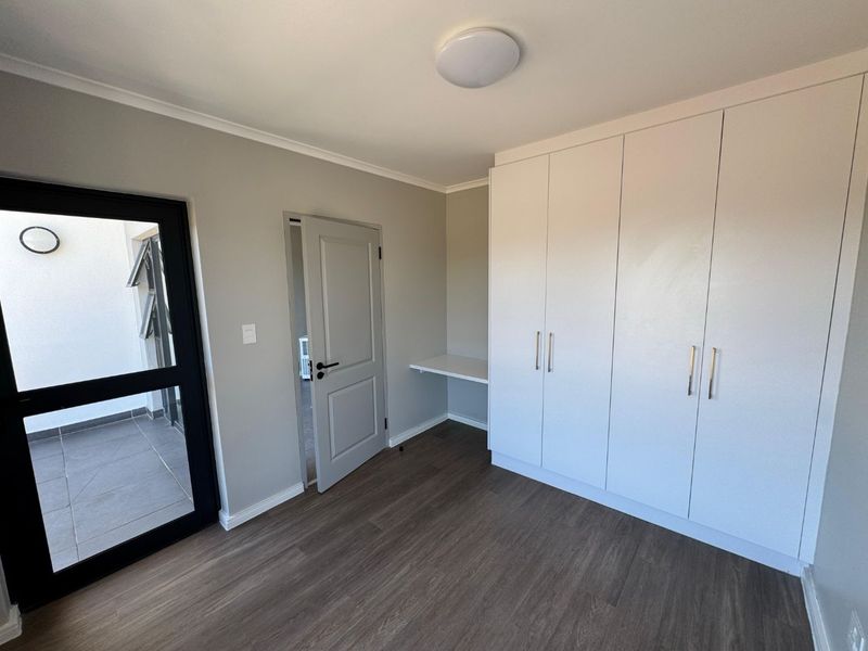 A bright, modern bedroom featuring neutral walls, laminate flooring, ample built-in cupboard space, a built-in desk, and direct access to an outdoor balcony area.