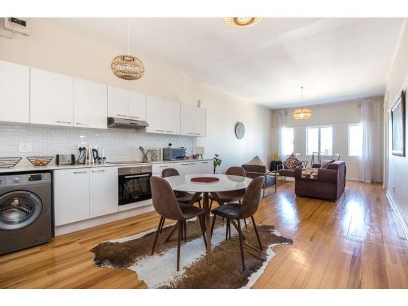 Bright open-plan living area featuring a modern kitchen with white cabinetry, subway tile backsplash, and integrated appliances, flowing into a spacious lounge with hardwood floors.