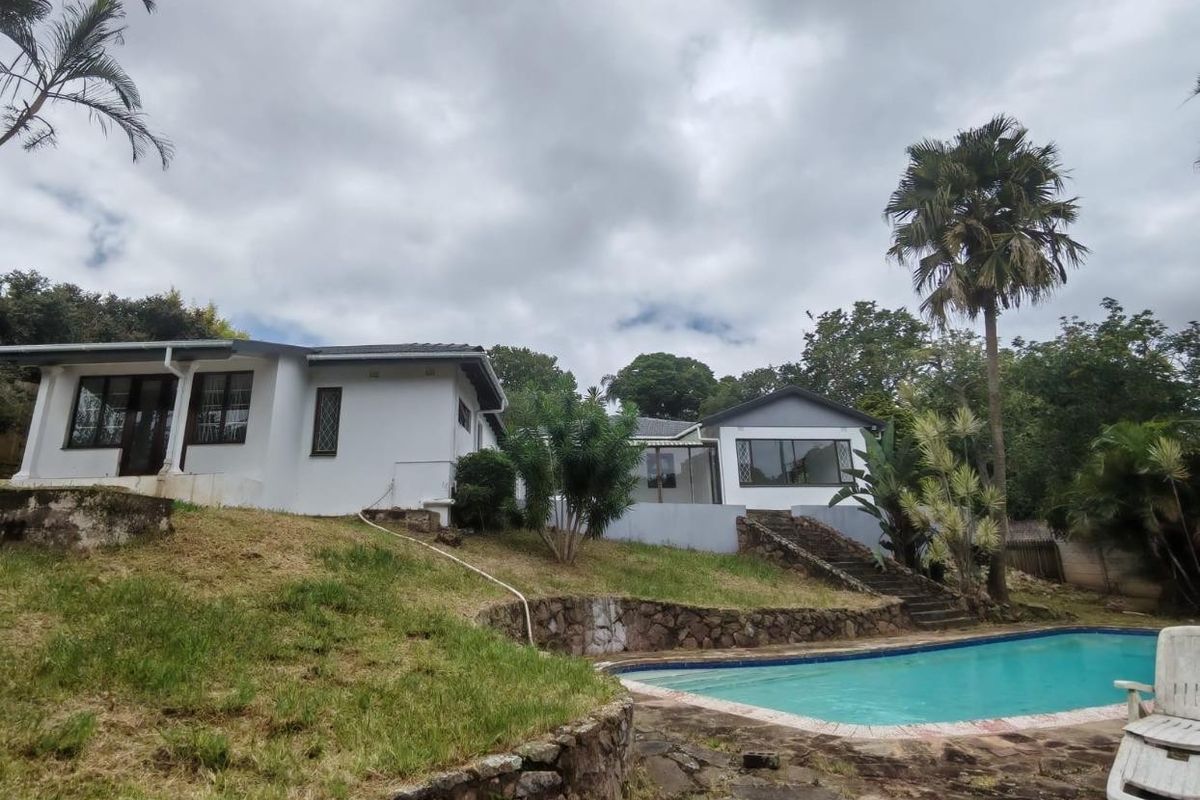 Rear exterior view of the house featuring a swimming pool, stone retaining walls, and a paved patio area set within a landscaped garden.