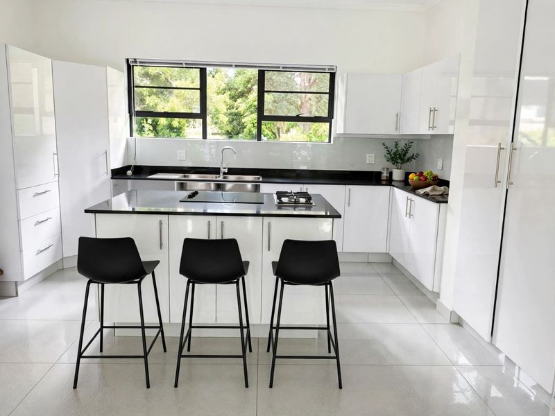 Modern kitchen featuring glossy white cabinetry, black stone countertops, a central island with a built-in hob, and large windows providing natural light.