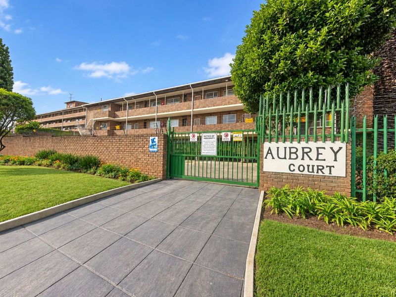 Front entrance view of Aubrey Court apartment complex featuring a paved driveway, secure metal gate, and brick boundary wall with landscaped garden.