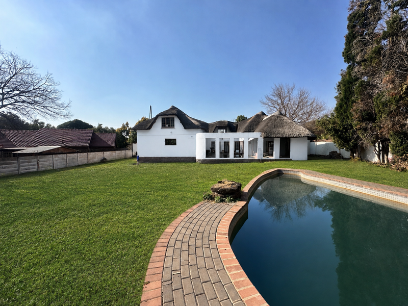 Rear view of a thatched-roof house featuring a large lawn, swimming pool with brick coping, and a covered patio area.