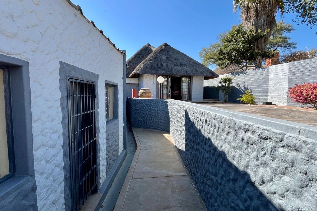 A paved walkway leads between two structures, featuring textured masonry walls and a traditional thatched-roof building in the background.