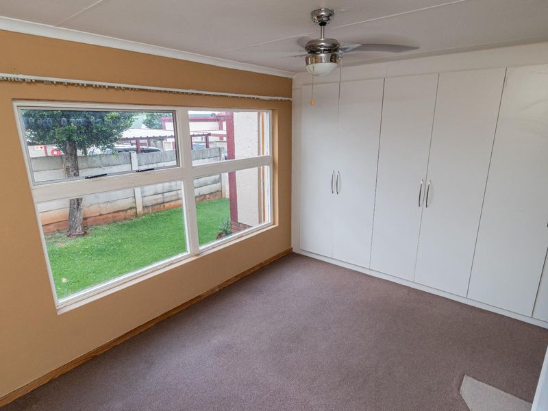 A bright, empty bedroom featuring built-in cupboards, a ceiling fan, and large windows overlooking the garden area.