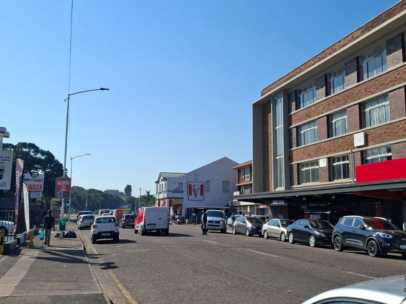 A street-level view of a busy commercial area featuring retail shopfronts, paved sidewalks, and urban infrastructure.