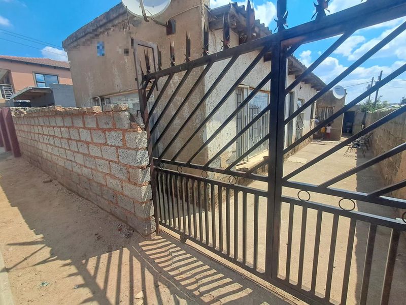 Front exterior view of a residential property featuring a metal security gate, concrete block boundary wall, and weathered building facade.