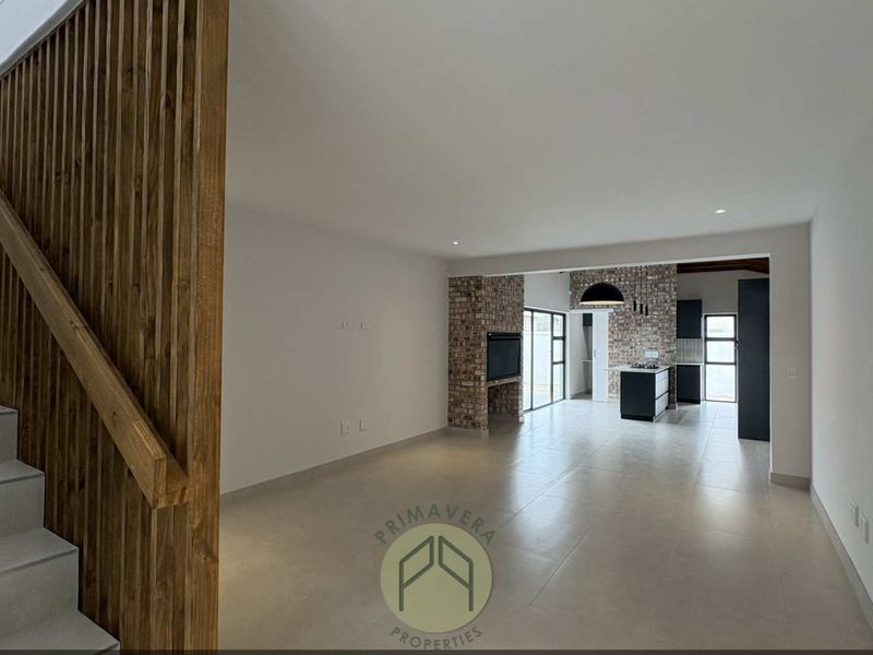 Spacious open-plan living and kitchen area featuring modern tiled floors, a striking timber staircase balustrade, and an exposed brick fireplace.