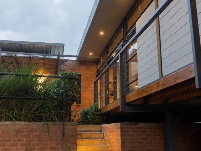 A modern rear exterior view featuring a brick retaining wall, concrete staircase leading to a timber deck, and large glass windows with integrated lighting.