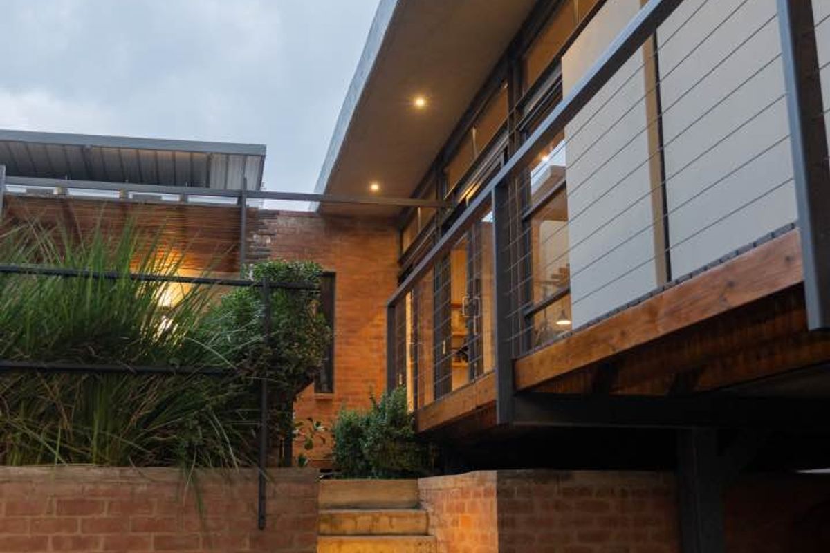 A modern rear exterior view featuring a brick retaining wall, concrete staircase leading to a timber deck, and large glass windows with integrated lighting.