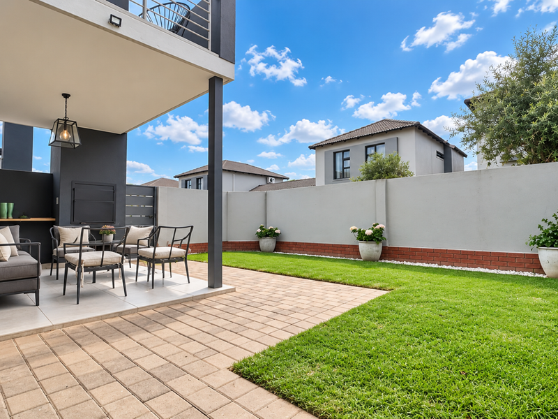 A well-maintained rear garden featuring a paved patio, covered entertainment area with a built-in braai, and a neat lawn enclosed by boundary walls.