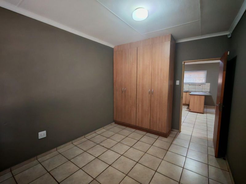 A clean, empty bedroom featuring tiled flooring and a built-in wooden wardrobe, with a view into the adjacent kitchen area.
