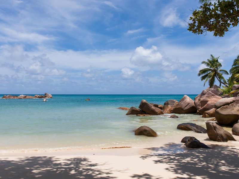 A scenic view of a pristine sandy beach with large granite boulders, tropical palm trees, and clear turquoise ocean waters.