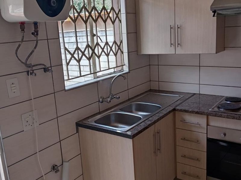 Compact kitchen featuring light wood cabinetry, built-in oven and hob, double sink, and wall-mounted electric water heater with tiled finishes.