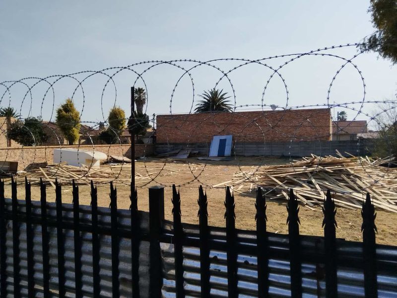 Vacant residential land enclosed by a metal fence with razor wire and a brick boundary wall, currently containing construction debris.