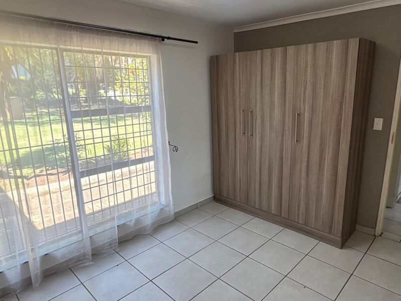 A bright, empty bedroom featuring tiled flooring, a built-in wooden wardrobe, and a sliding glass door with security bars leading to the garden.