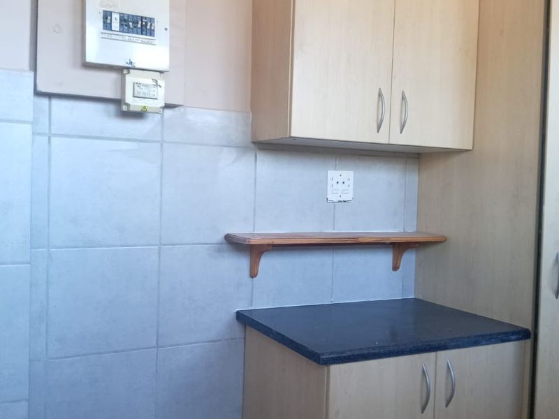 A compact kitchen area featuring light wood-finish cabinetry, dark laminate countertops, and a tiled splashback, with an exposed electrical distribution board on the wall.