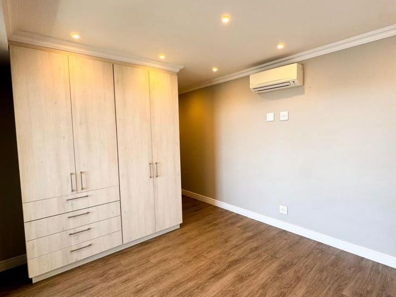 A bright, empty bedroom featuring light wood-finish built-in cupboards, laminate flooring, recessed ceiling lighting, and a wall-mounted air conditioning unit.