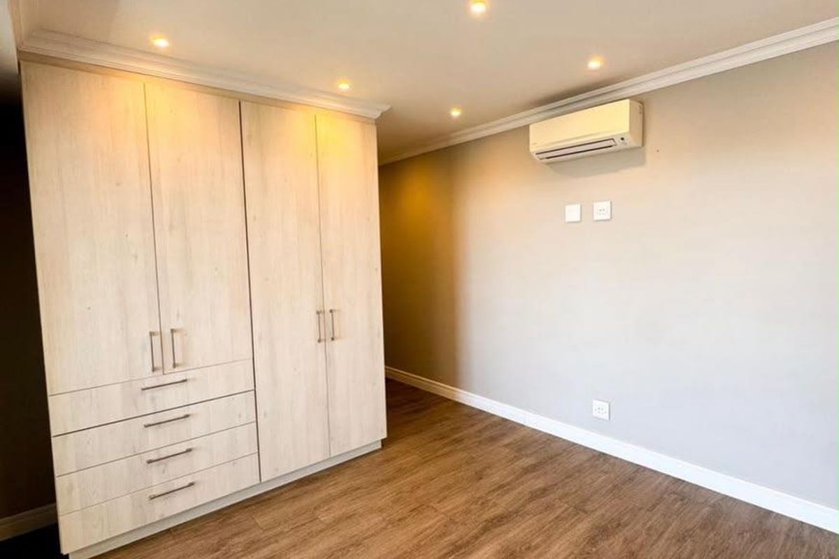 A bright, empty bedroom featuring light wood-finish built-in cupboards, laminate flooring, recessed ceiling lighting, and a wall-mounted air conditioning unit.