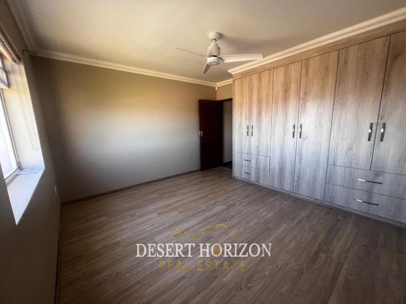 A spacious, empty bedroom featuring modern wood-look laminate flooring, extensive built-in cupboards, and a ceiling fan.