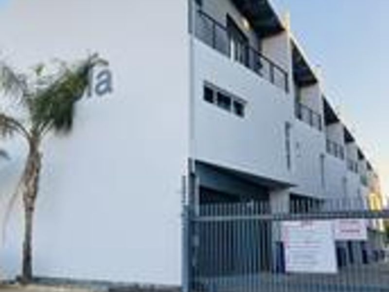 Modern multi-story apartment complex exterior featuring clean white facades, private balconies, and secure gated access with paved driveway.